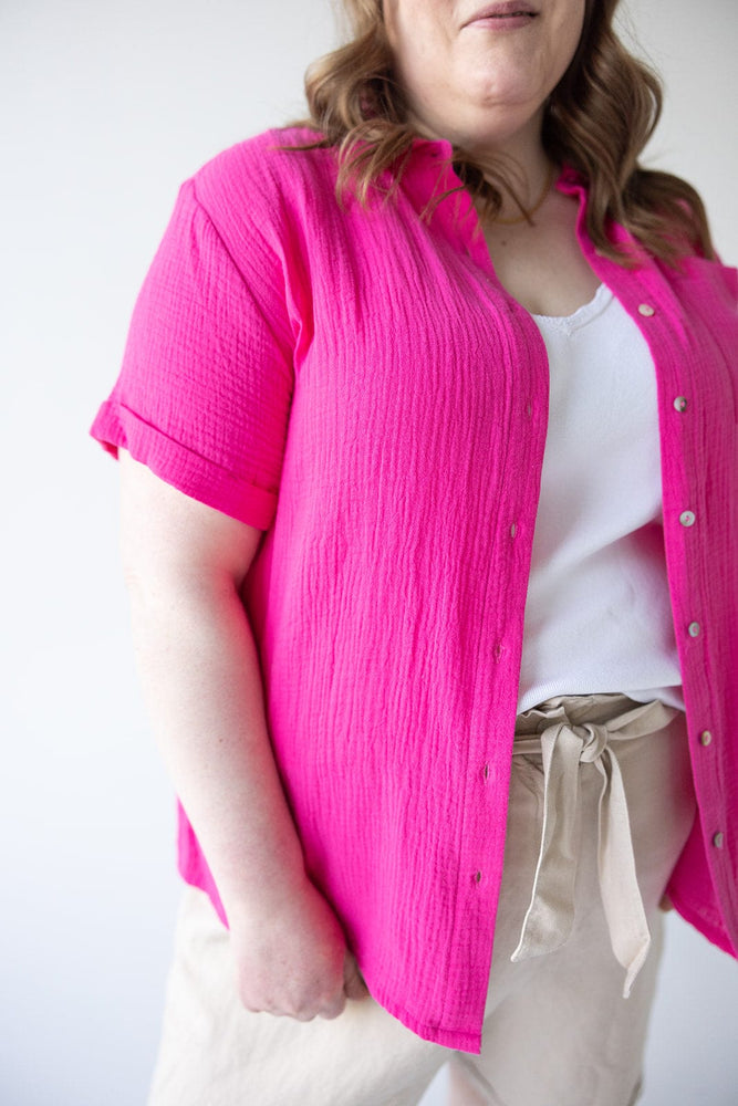 A person wearing a vibrant TEXTURED COTTON BUTTON UP BLOUSE IN AZALEA by Yesta over a white top, beige pants with a tied waistband, and having wavy hair.