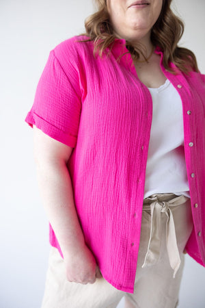 A person wearing a vibrant TEXTURED COTTON BUTTON UP BLOUSE IN AZALEA by Yesta over a white top, beige pants with a tied waistband, and having wavy hair.