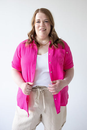 A person with shoulder-length hair wearing a bright pink Yesta Textured Cotton Button-Up Blouse in Azalea, a white top, and beige pants stands smiling against a plain background.