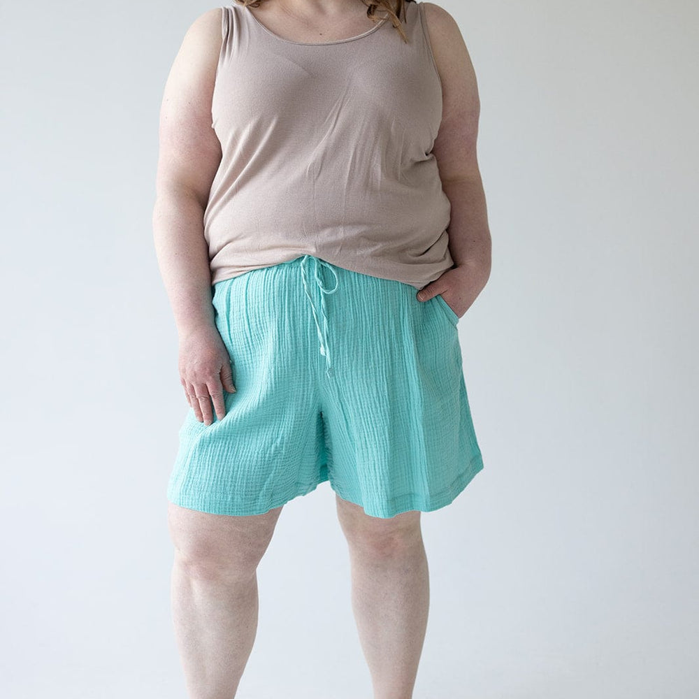 A person stands against a plain background wearing a beige tank top, Maui Textured Cotton Shorts with an elastic waistband, and brown sandals.