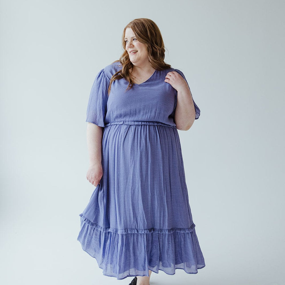 A woman stands against a plain background, wearing the Mikarose Textured Flutter Sleeve Dress with Kick Hem in Ultra Purple and black flats. Her left hand touches her hair as she has a slight smile on her face.