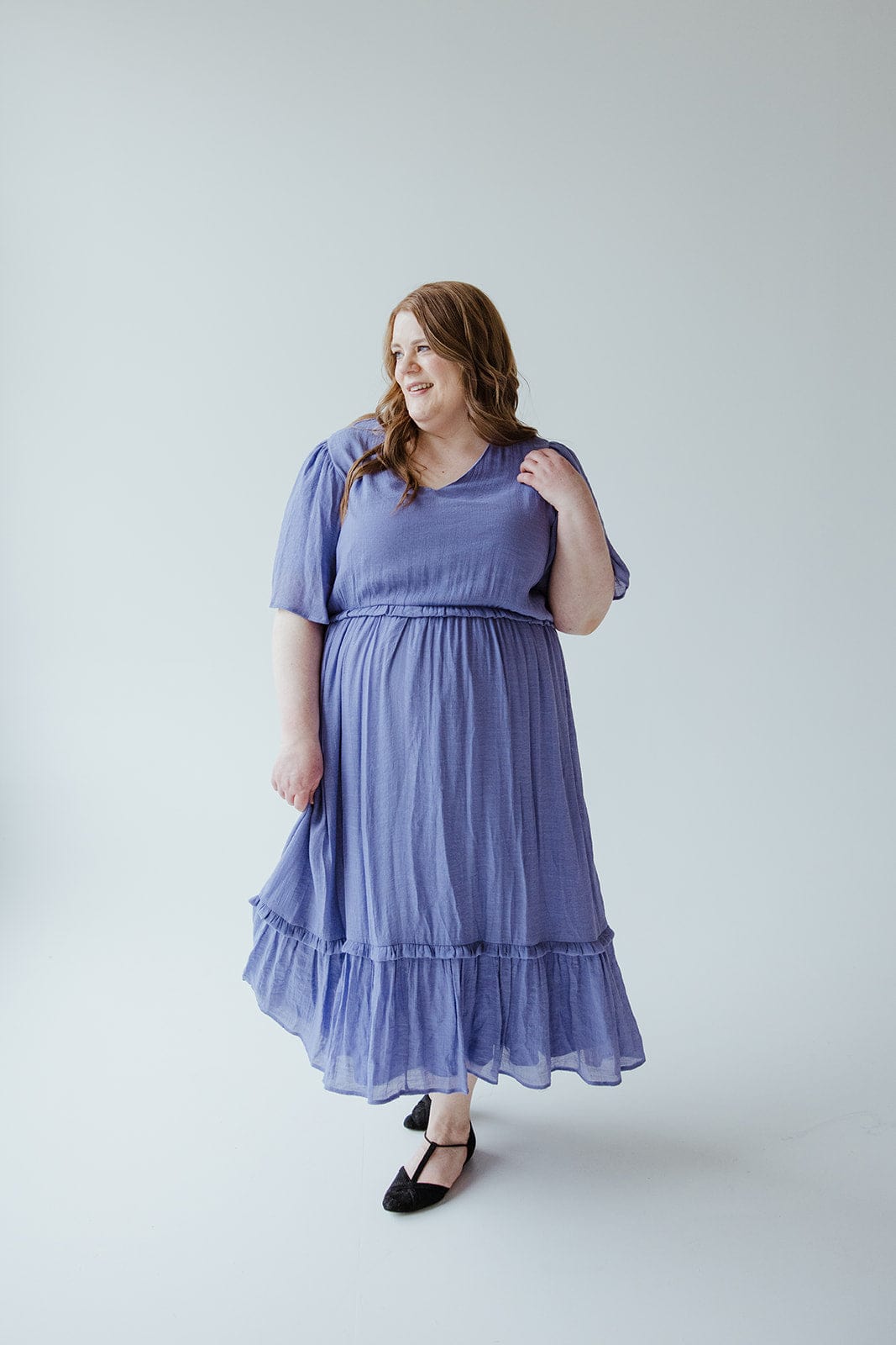 A woman stands against a plain background, wearing the Mikarose Textured Flutter Sleeve Dress with Kick Hem in Ultra Purple and black flats. Her left hand touches her hair as she has a slight smile on her face.