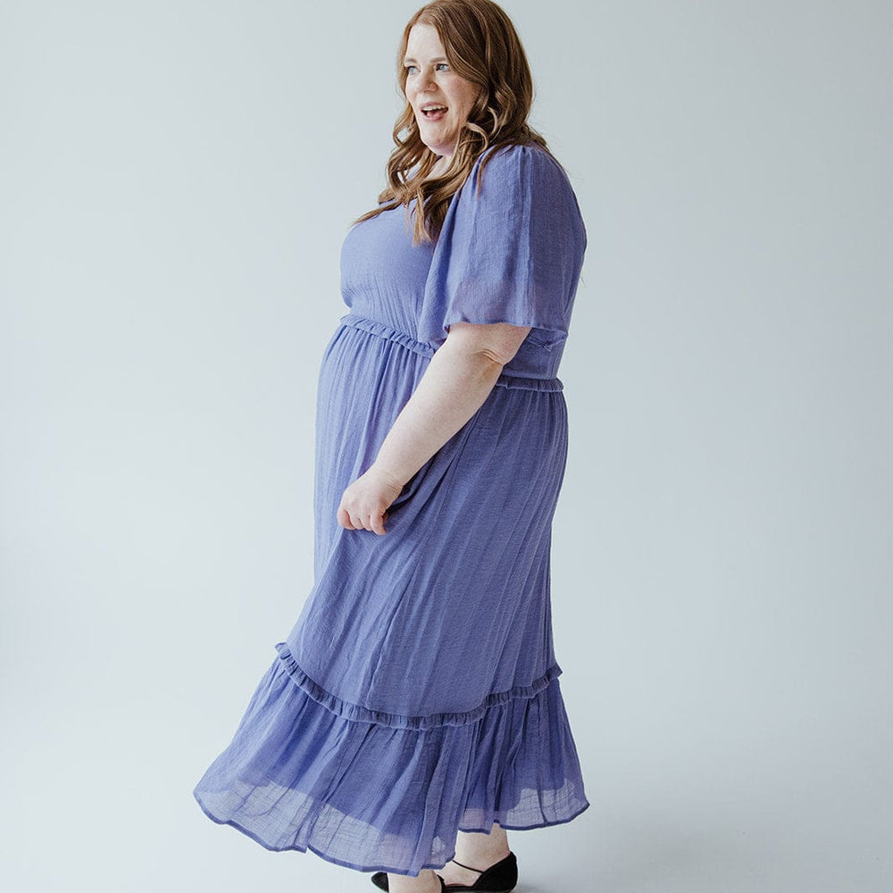 A woman in a Mikarose TEXTURED FLUTTER SLEEVE DRESS WITH KICK HEM IN ULTRA PURPLE and black shoes stands against a light gray background, smiling and looking to the side.