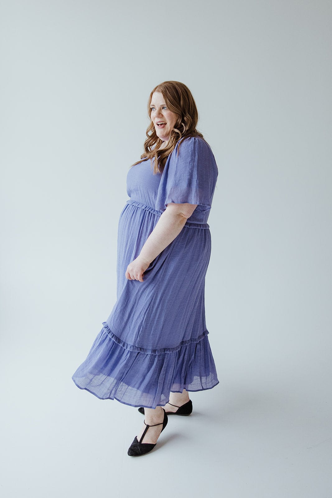 A woman in a Mikarose TEXTURED FLUTTER SLEEVE DRESS WITH KICK HEM IN ULTRA PURPLE and black shoes stands against a light gray background, smiling and looking to the side.