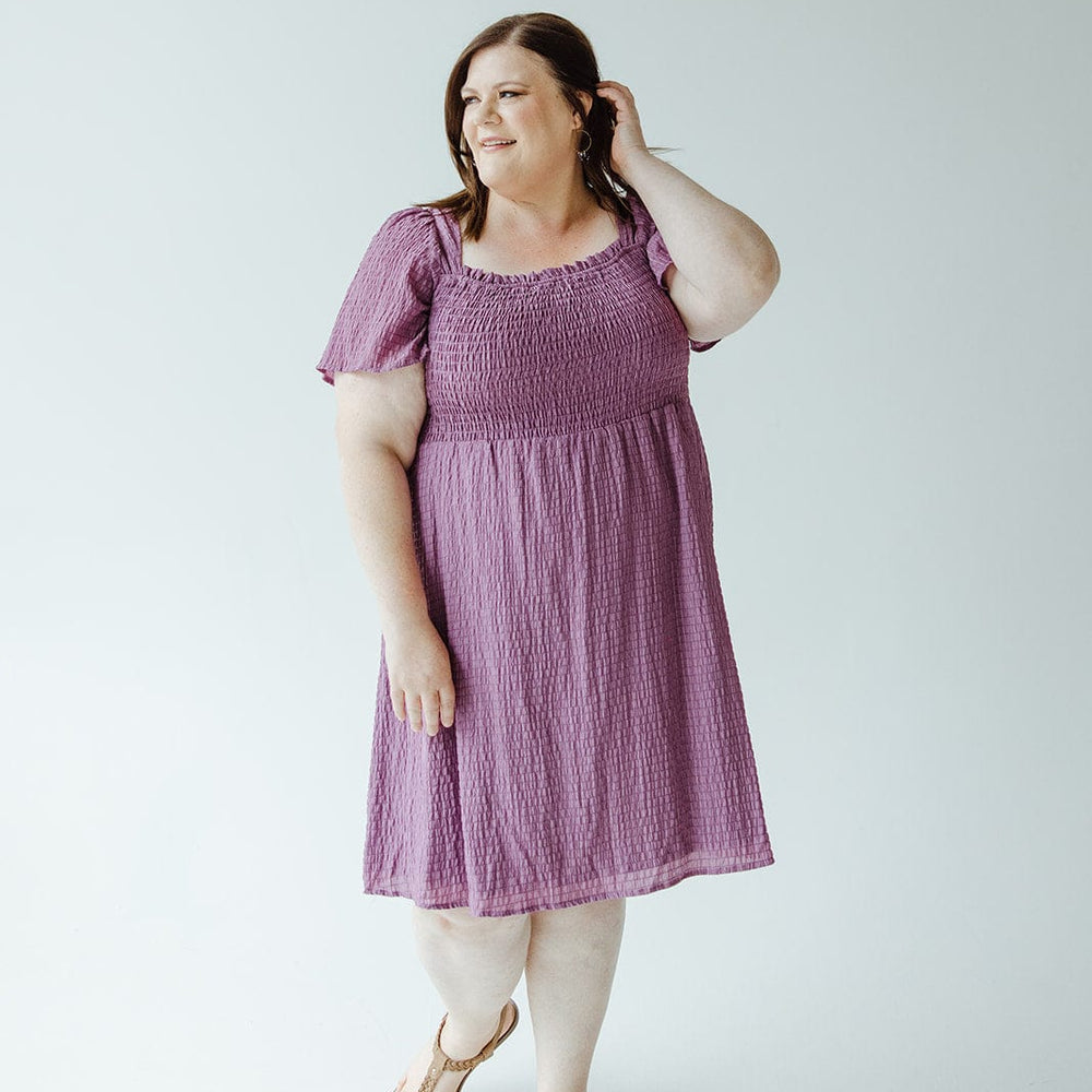 A woman in a Mikarose TEXTURED KNEE LENGTH DRESS IN DEWBERRY poses against a white background, with one hand touching her hair and the other resting at her side. She is wearing sandals.