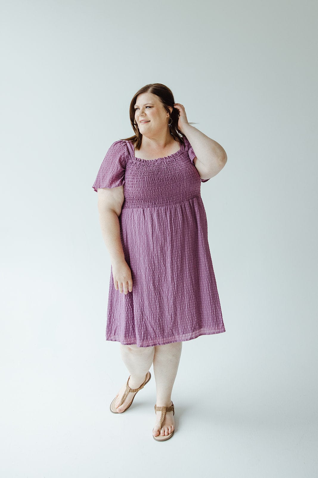 A woman in a Mikarose TEXTURED KNEE LENGTH DRESS IN DEWBERRY poses against a white background, with one hand touching her hair and the other resting at her side. She is wearing sandals.