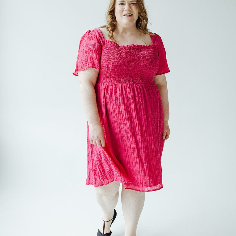 A woman wearing a TEXTURED KNEE LENGTH DRESS IN VIVA MAGENTA by Mikarose, with a smocked bodice and black shoes, stands on a plain white background.