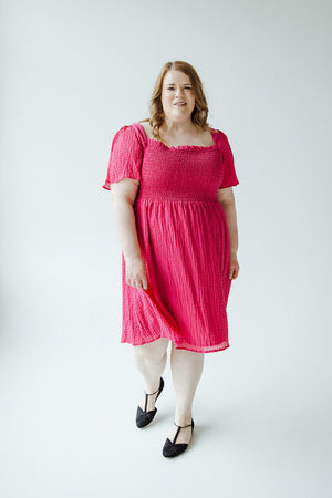 A woman wearing a TEXTURED KNEE LENGTH DRESS IN VIVA MAGENTA by Mikarose, with a smocked bodice and black shoes, stands on a plain white background.