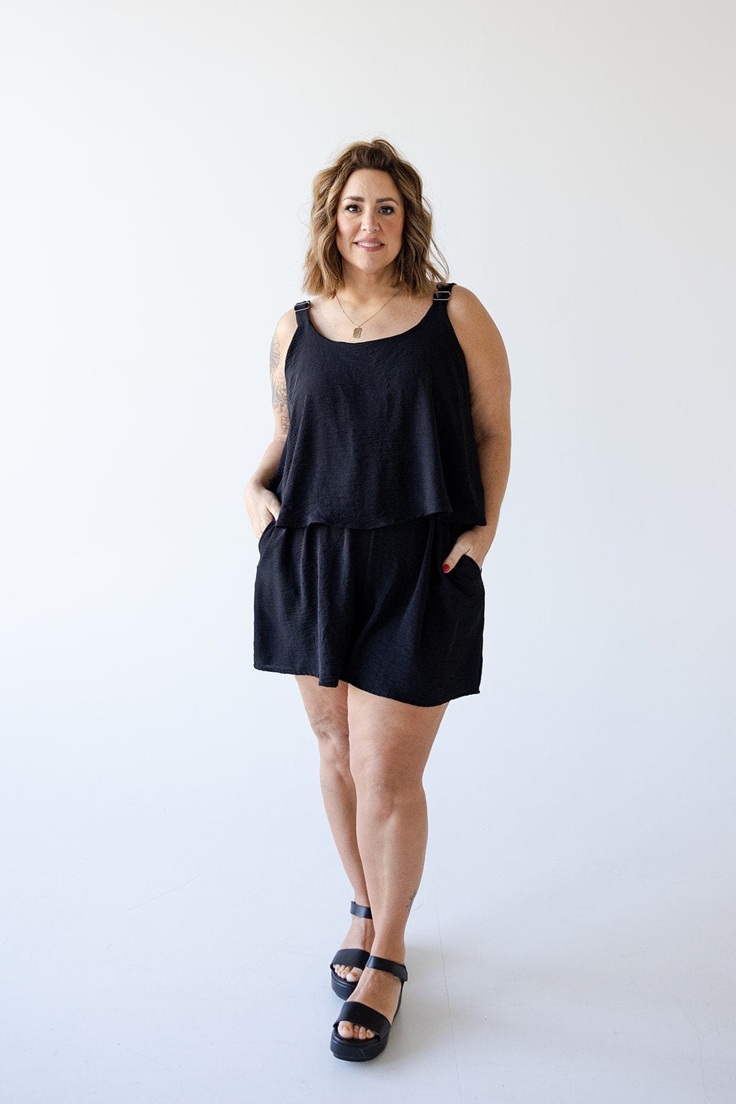 A person stands in front of a plain white background, showcasing summer fashion in a lightweight romper with adjustable straps. The ensemble, featuring a black sleeveless top and shorts, is completed with hands casually placed in pockets and black sandals.