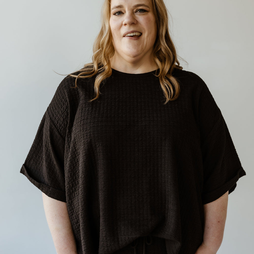 A person with long hair stands against a plain white background, wearing Mikarose's textured oversized tee with rolled cuffs and a scooped neckline, along with matching pants.