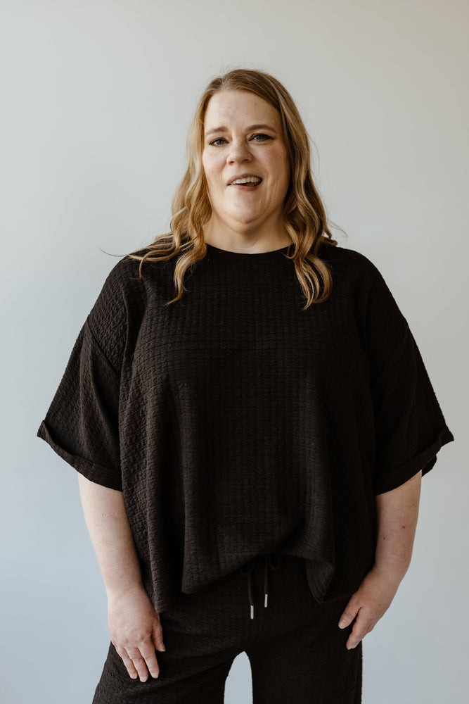 A person with long hair stands against a plain white background, wearing Mikarose's textured oversized tee with rolled cuffs and a scooped neckline, along with matching pants.