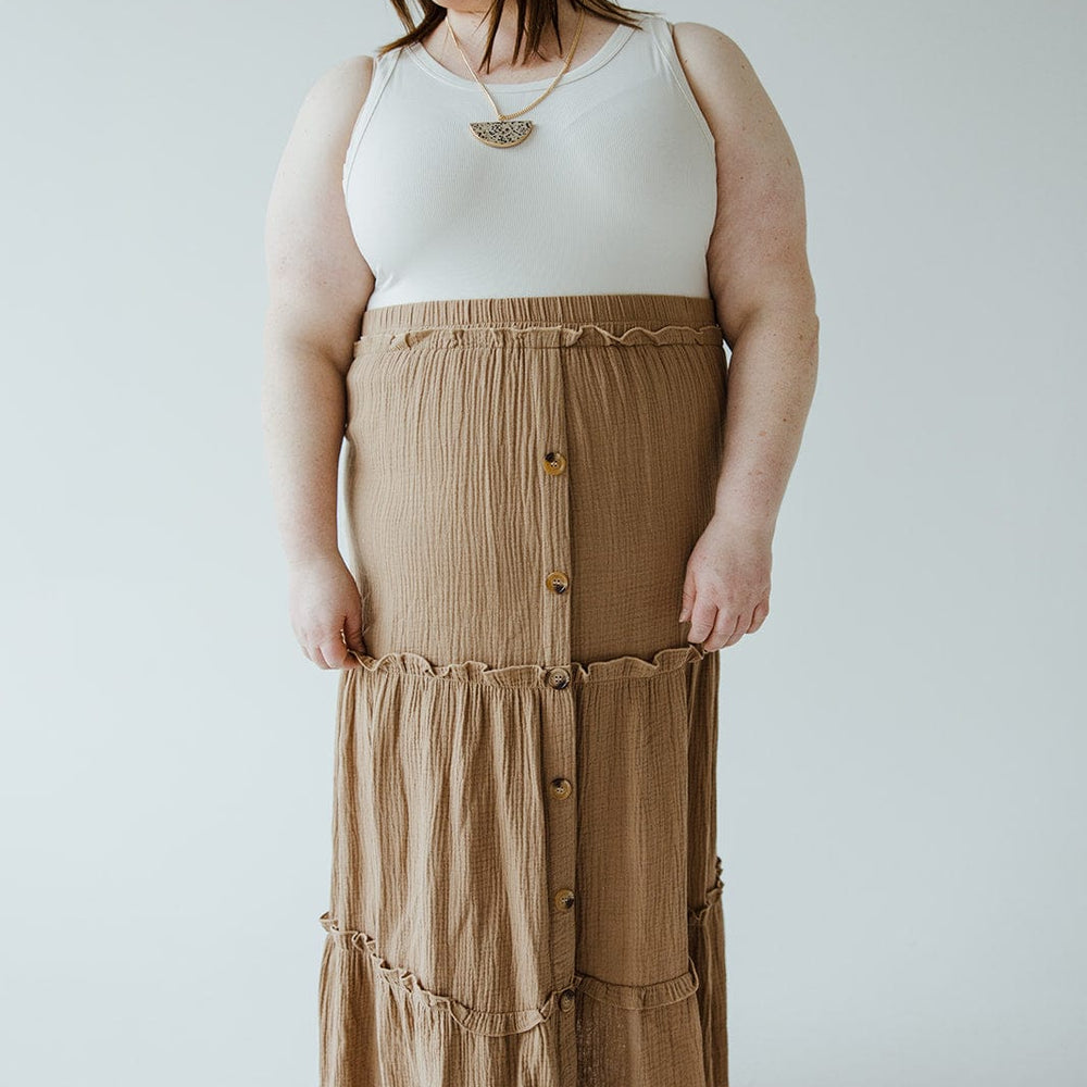 A person stands against a plain background wearing a white sleeveless top and a long brown Mikarose TEXTURED TIERED SKIRT WITH BUTTONS, perfect for a summer wardrobe.