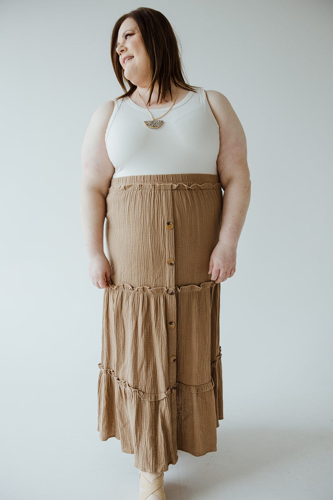 A person stands against a plain background wearing a white sleeveless top and a long brown Mikarose TEXTURED TIERED SKIRT WITH BUTTONS, perfect for a summer wardrobe.