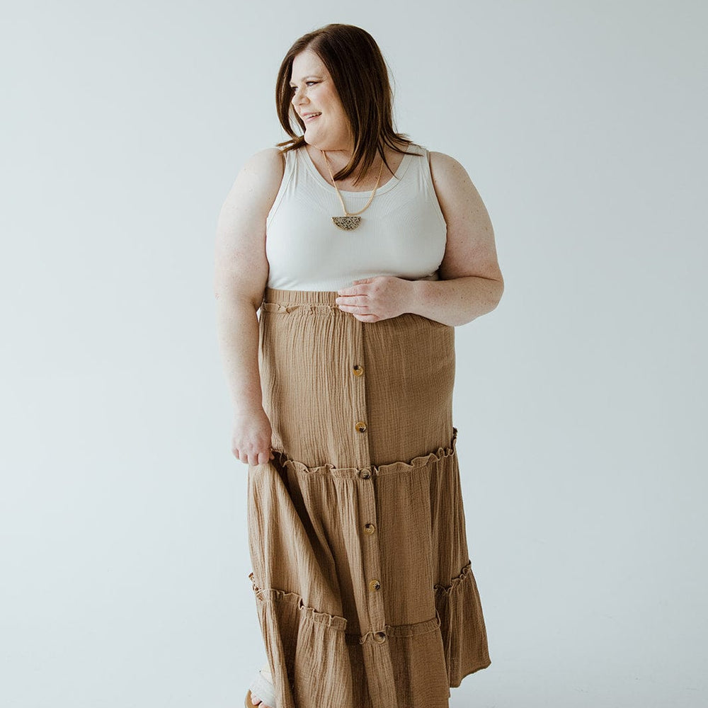 A woman standing against a plain white background, wearing a white sleeveless top and a long Mikarose TEXTURED TIERED SKIRT WITH BUTTONS with an elastic waistband, looking to her left with a slight smile. Perfect for any summer wardrobe.