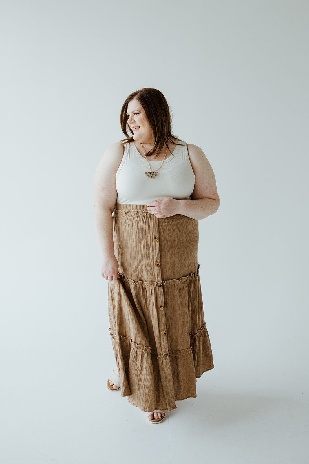 A woman standing against a plain white background, wearing a white sleeveless top and a long Mikarose TEXTURED TIERED SKIRT WITH BUTTONS with an elastic waistband, looking to her left with a slight smile. Perfect for any summer wardrobe.