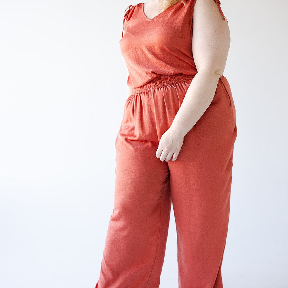 A woman wearing Love Marlow's Textured Wide Leg Satin Pants in Copper stands and smiles, glancing off to the side. She pairs the loose-fitting pants, which feature an elastic waistband for comfort, with a sleeveless top. Her long hair flows down as she completes her look with black sandals.