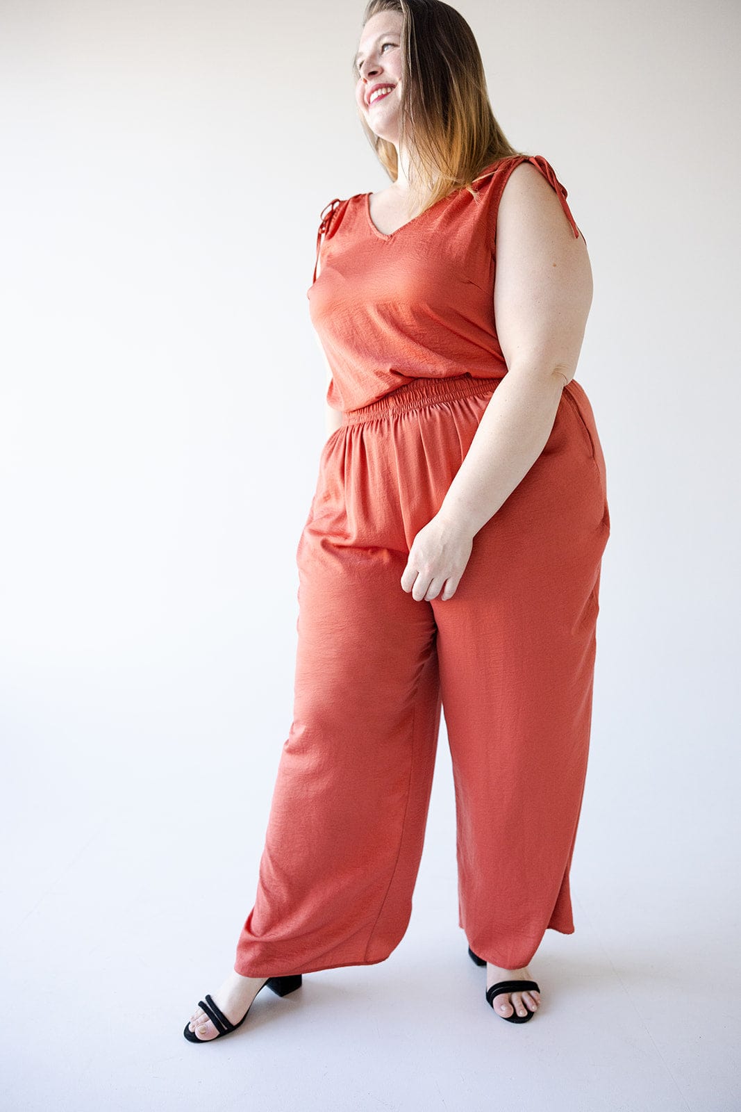 A woman wearing Love Marlow's Textured Wide Leg Satin Pants in Copper stands and smiles, glancing off to the side. She pairs the loose-fitting pants, which feature an elastic waistband for comfort, with a sleeveless top. Her long hair flows down as she completes her look with black sandals.