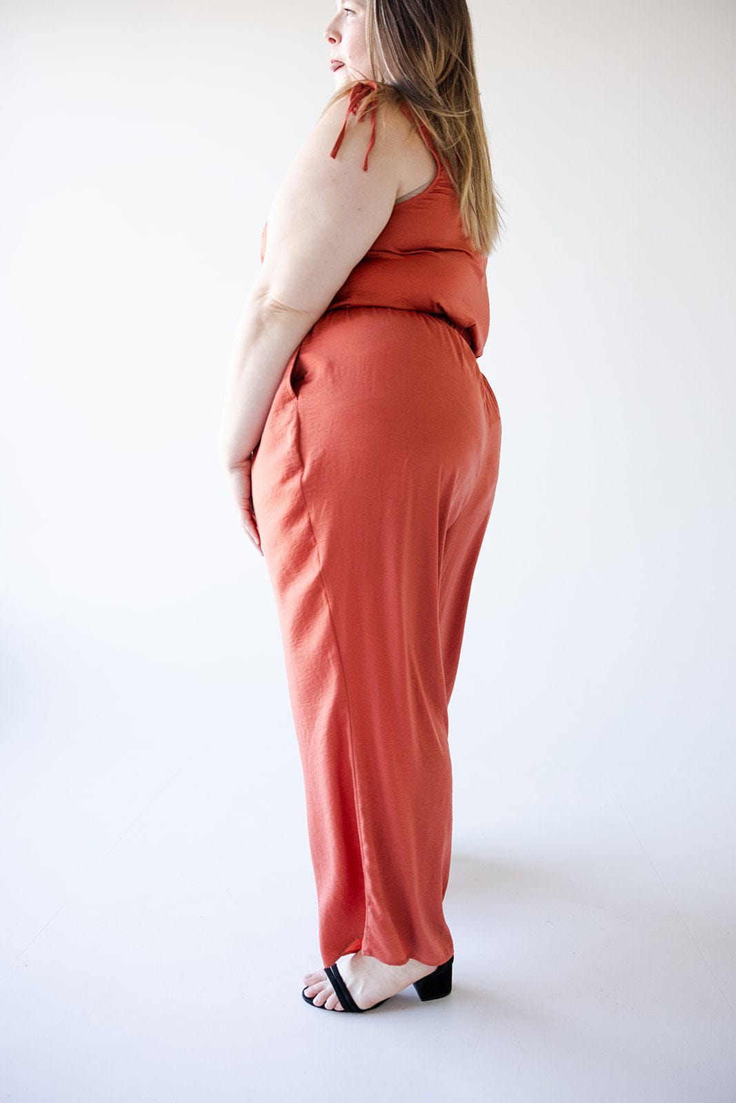 A person wearing Textured Wide Leg Satin Pants in Copper from Love Marlow, paired with black sandals, stands sideways in a minimalist setting.