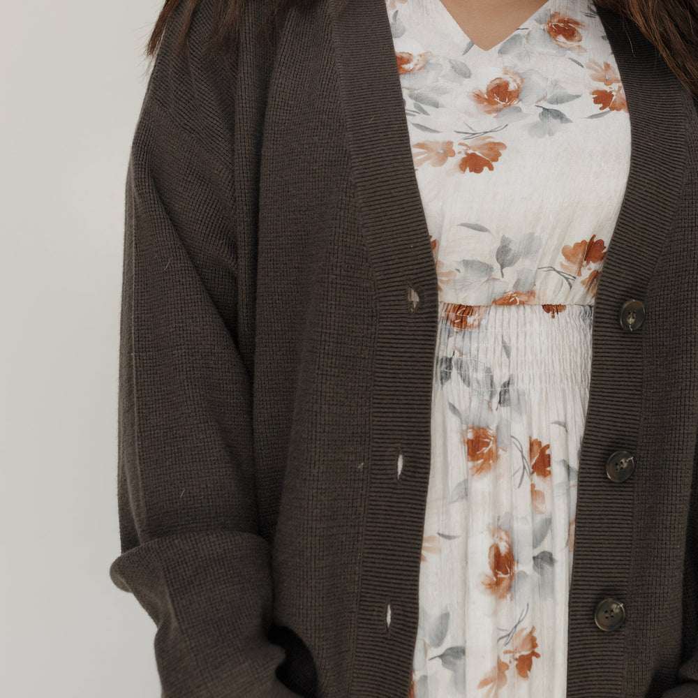 Person wearing a brown cardigan over a floral dress against a plain background