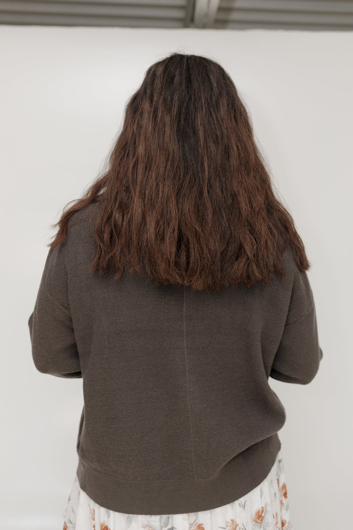 Person wearing a brown sweater with long hair, standing against a white wall.