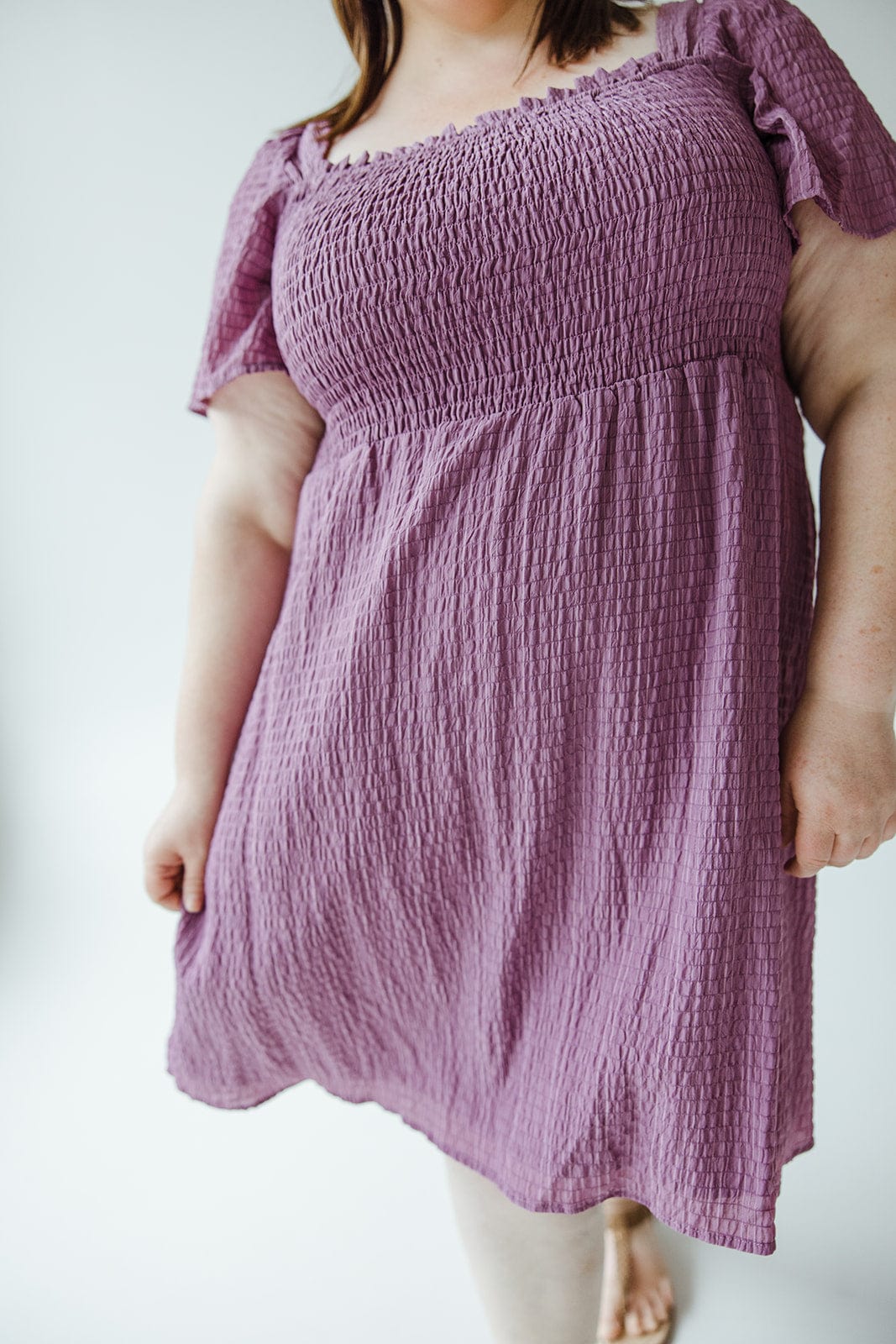 Person wearing a purple, short-sleeved TEXTURED KNEE LENGTH DRESS IN DEWBERRY by Mikarose with an a-line skirt, standing against a plain background.