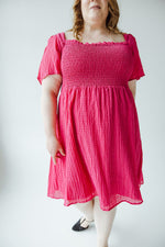 A person with long hair wearing a bright pink TEXTURED KNEE LENGTH DRESS IN VIVA MAGENTA by Mikarose and black shoes is standing and facing away from the camera against a plain white background.