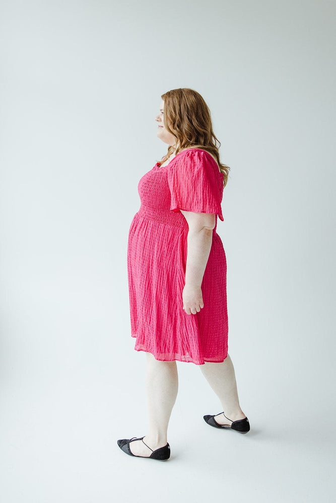 A woman with long hair, clad in a Mikarose TEXTURED KNEE LENGTH DRESS IN VIVA MAGENTA and black flats, walks against a plain, light-colored background.