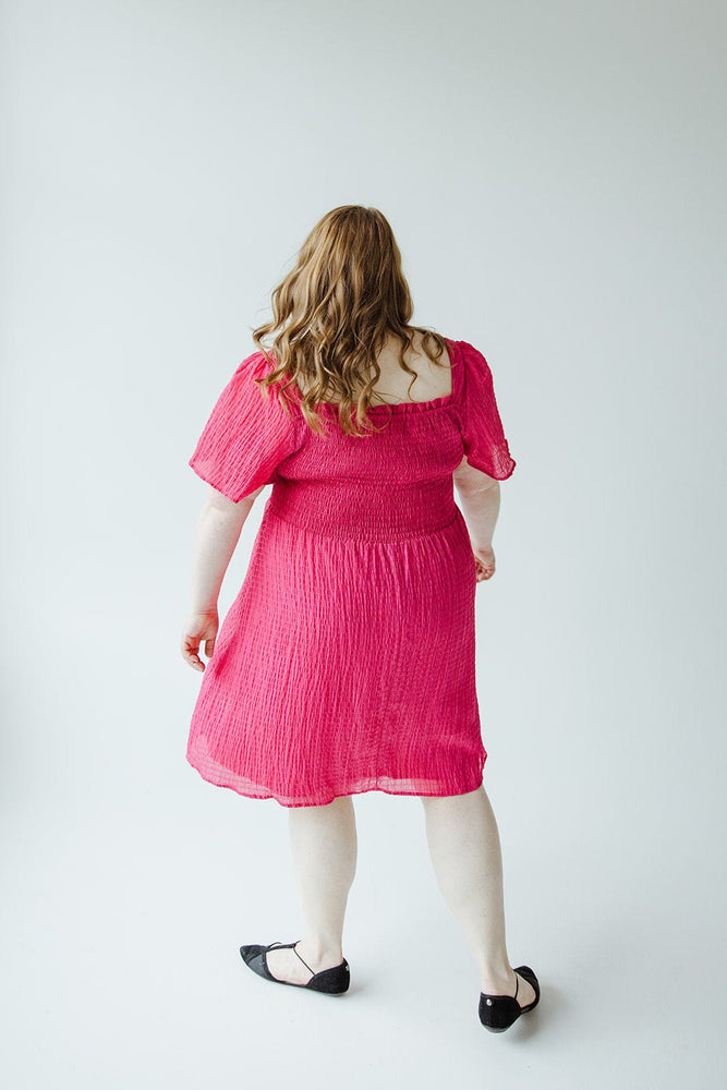 A person with long hair wearing a bright pink TEXTURED KNEE LENGTH DRESS IN VIVA MAGENTA by Mikarose and black shoes is standing and facing away from the camera against a plain white background.