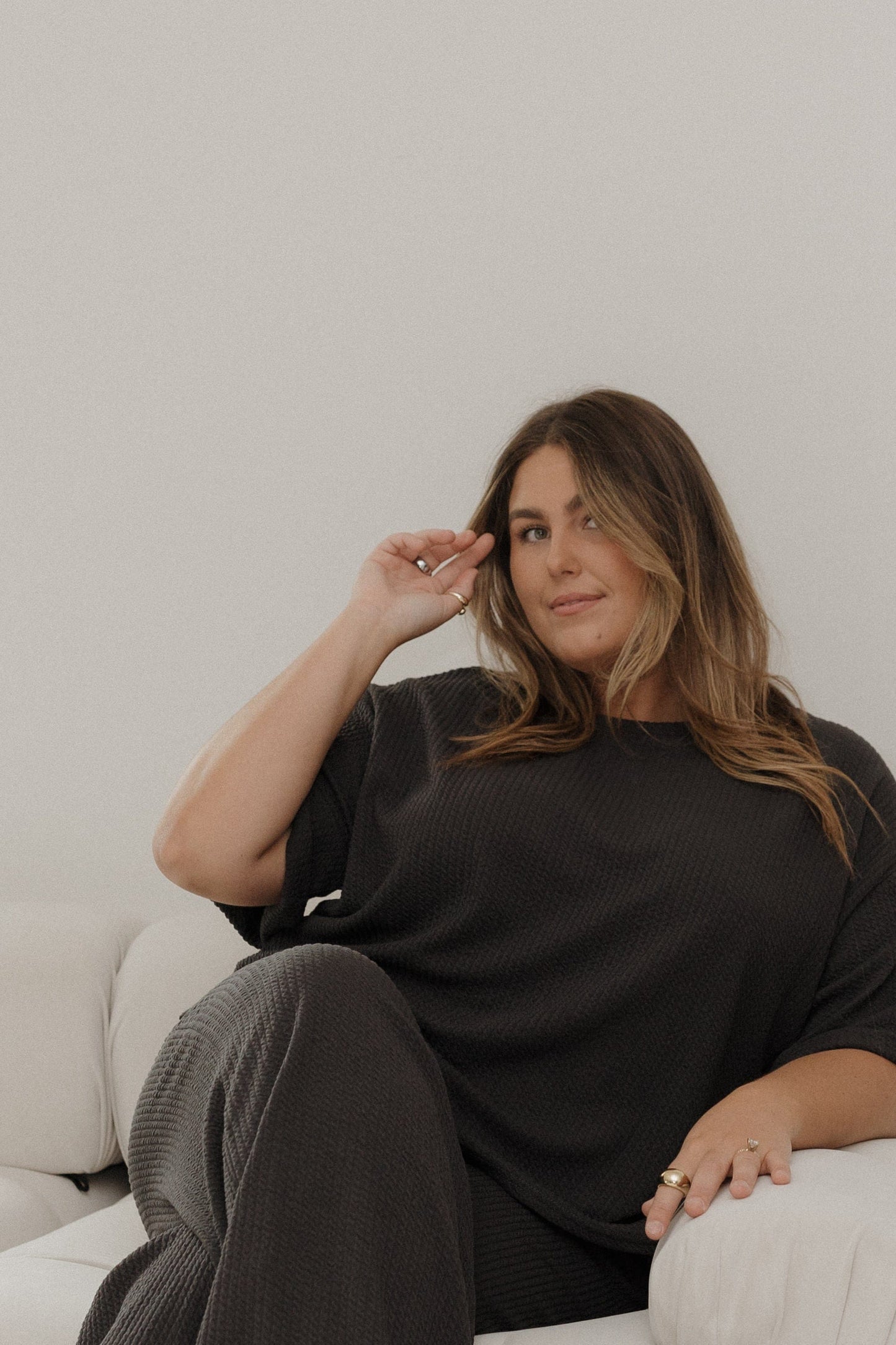 Woman sitting on a couch wearing a black sweater and gray pants, with a neutral background.