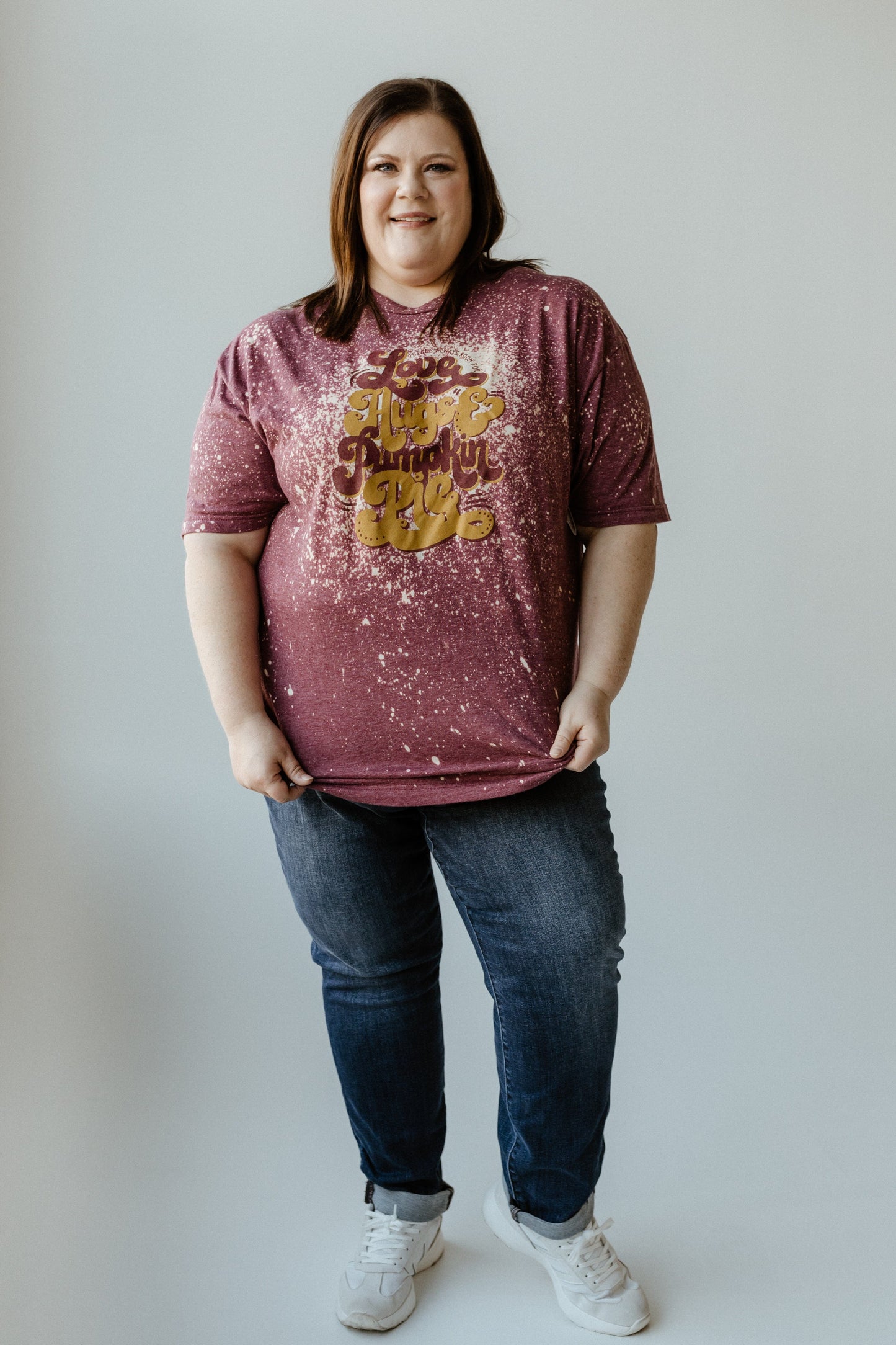 Person wearing a maroon speckled, hand-bleached T-shirt with "Love Peace Pumpkin Pie" text, blue jeans, and white sneakers, standing against a plain background.