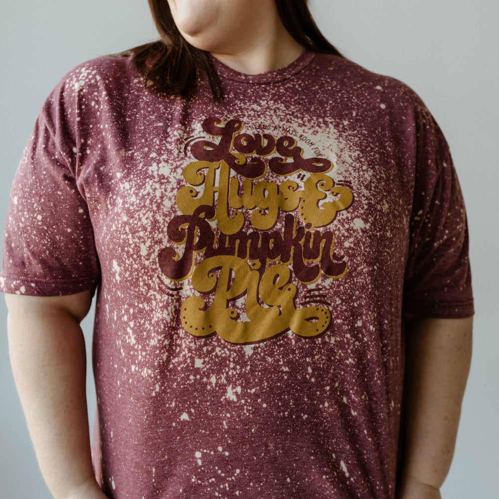 A person in a hand bleached maroon, splattered T-shirt with the text "Love, Hugs & Pumpkin Pie," reminiscent of Grandma's warmth, looks to the side and smiles.