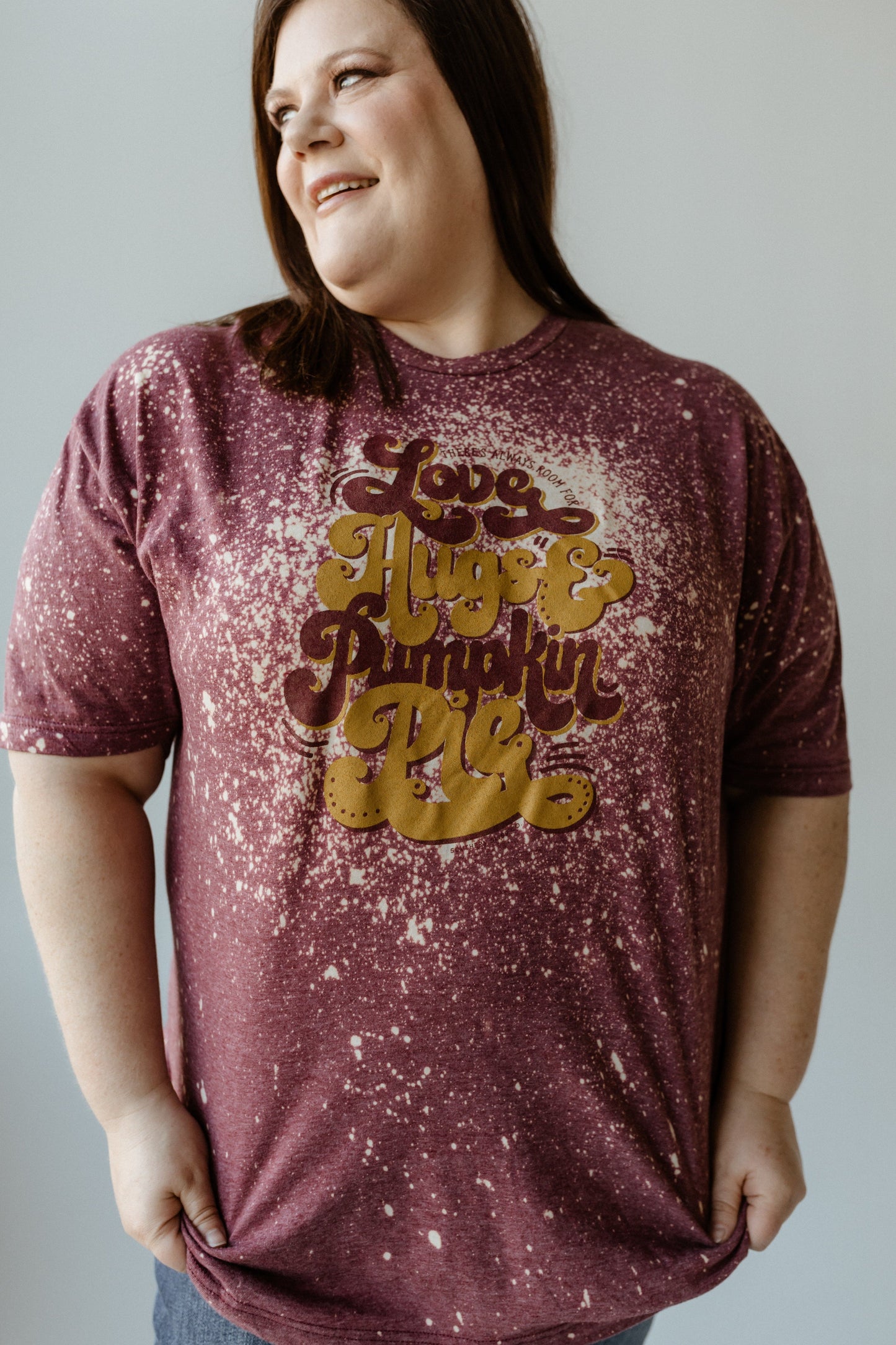 A person in a hand bleached maroon, splattered T-shirt with the text "Love, Hugs & Pumpkin Pie," reminiscent of Grandma's warmth, looks to the side and smiles.