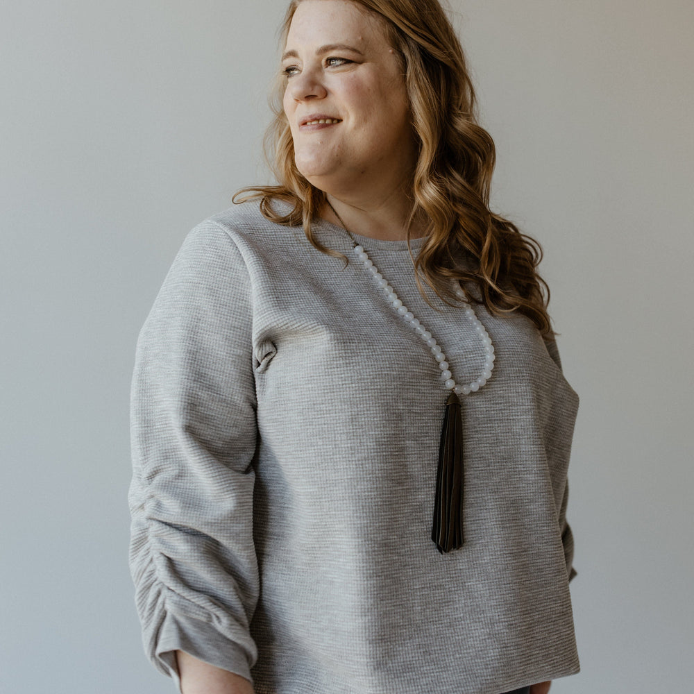 A woman with wavy hair in a light gray, silver lurex sweater and a long beaded necklace stands against a plain background, looking to her left.