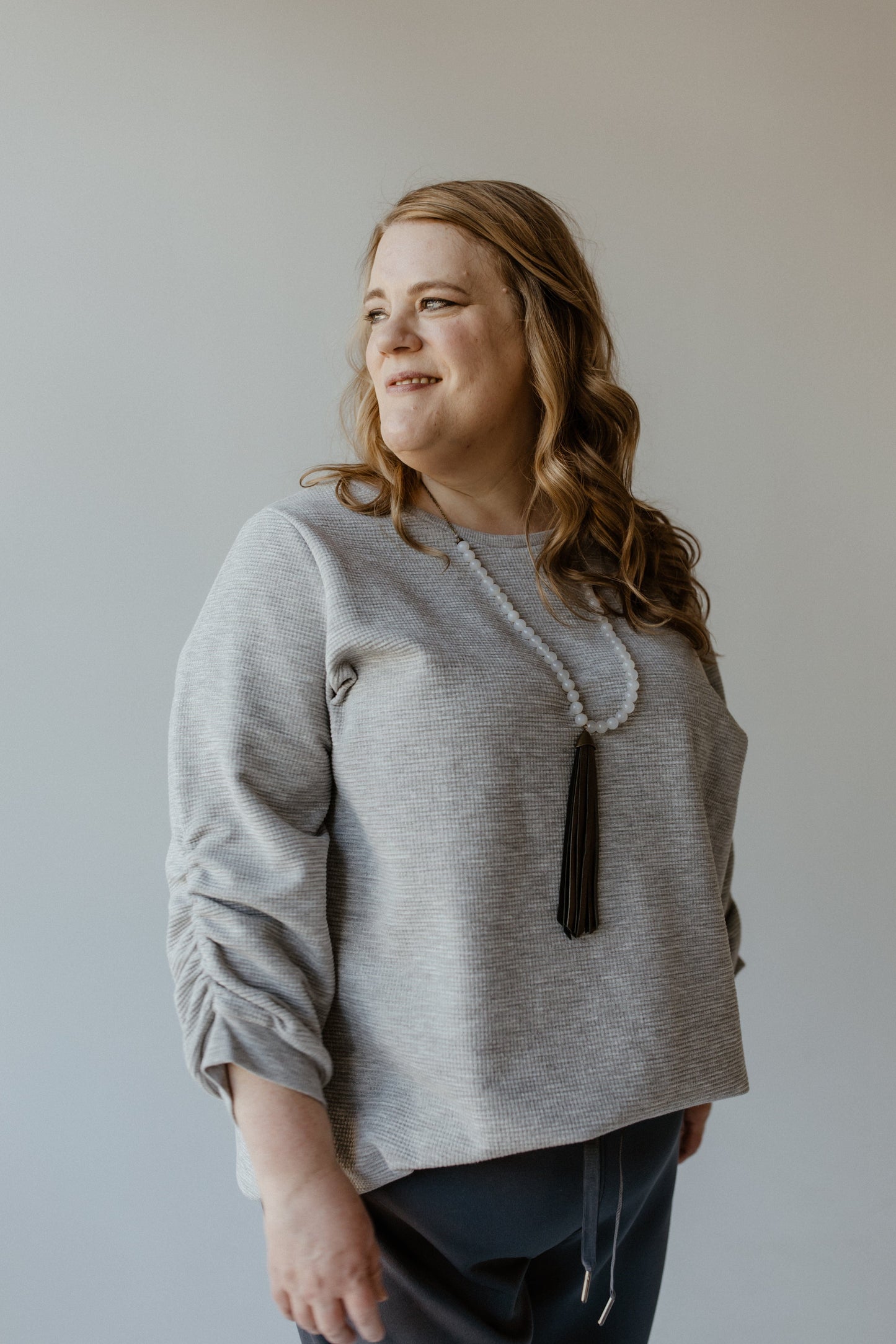 A woman with wavy hair in a light gray, silver lurex sweater and a long beaded necklace stands against a plain background, looking to her left.