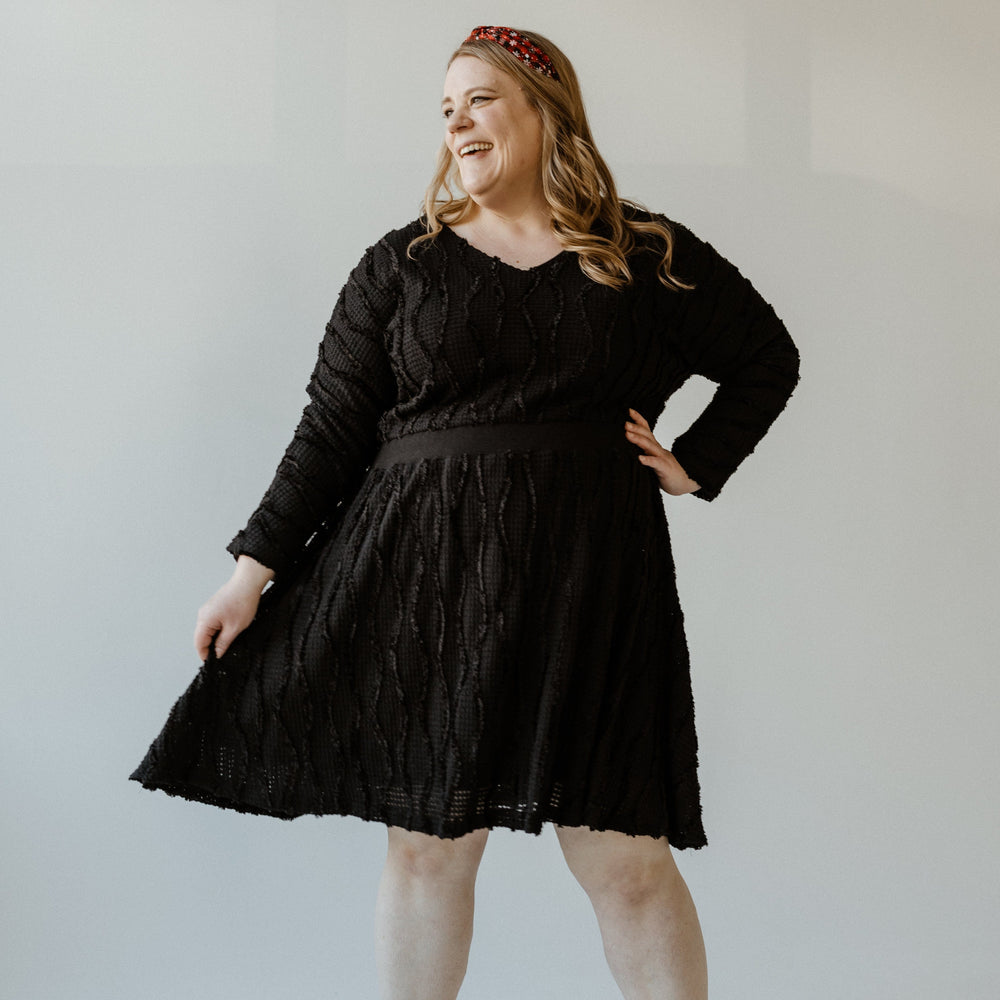 A person in a sleek black thermal texture dress and matching shoes is smiling while posing with one hand on their hip against a light background.