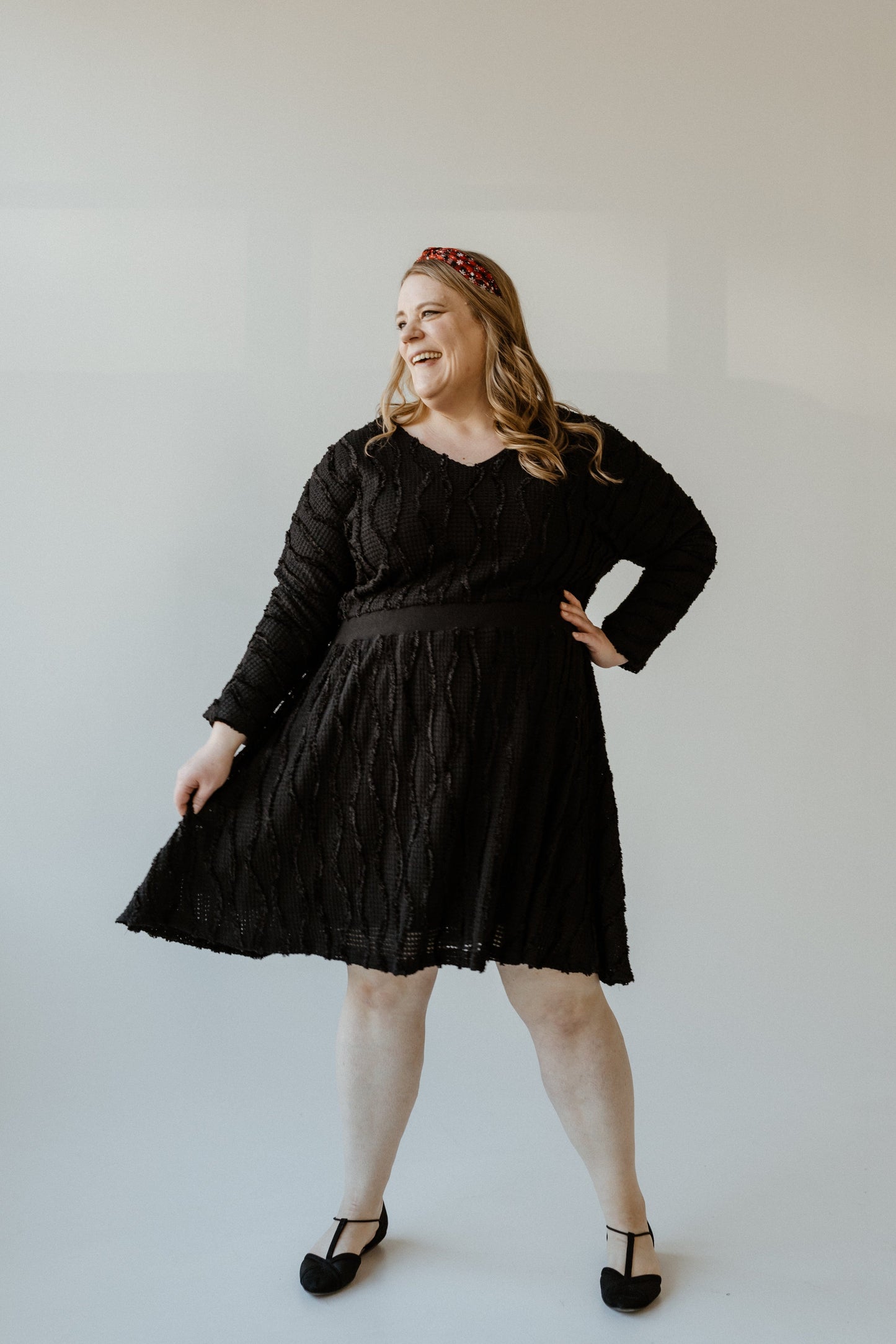 A person in a sleek black thermal texture dress and matching shoes is smiling while posing with one hand on their hip against a light background.