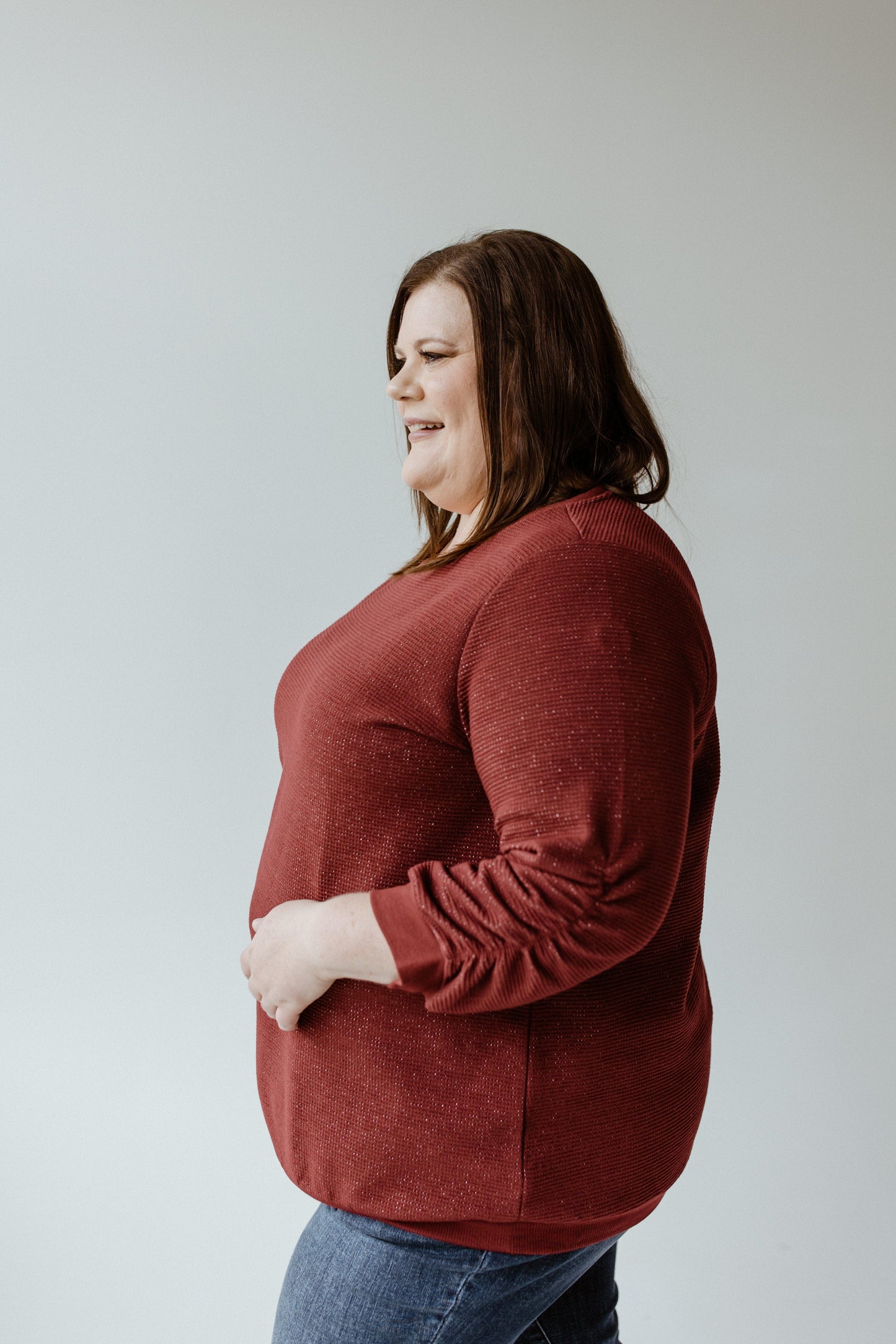 A person with long, brown hair wearing a maroon sweater and jeans stands against a plain background, smiling; their outfit radiates elegance with the THERMAL SPARKLE 3/4 TEE IN DARK CUT RUBY by Yesta offering a luxurious, personalized fit.