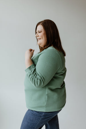 Wearing the Yesta Thermal Sparkle 3/4 Tee in Frosted Juniper paired with blue jeans, a woman is smiling with subtle silver accents shimmering as she looks to the side against a light background.