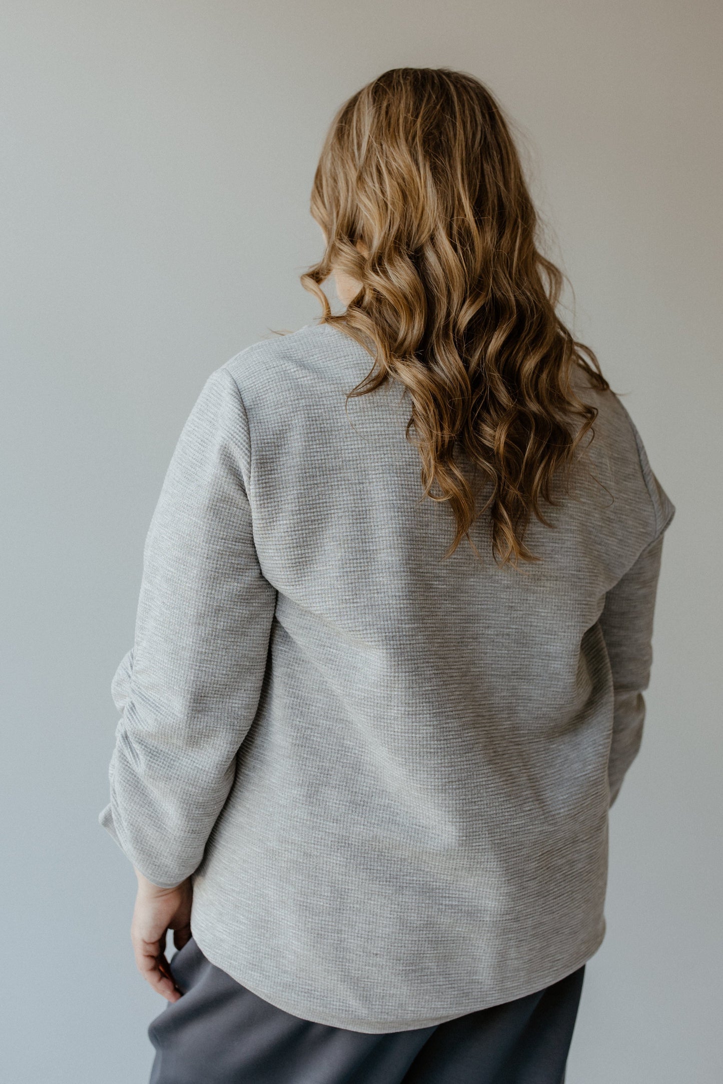 A person with long wavy hair is wearing a gray sweater with ruched sleeves and dark pants, facing away from the camera against a plain background.