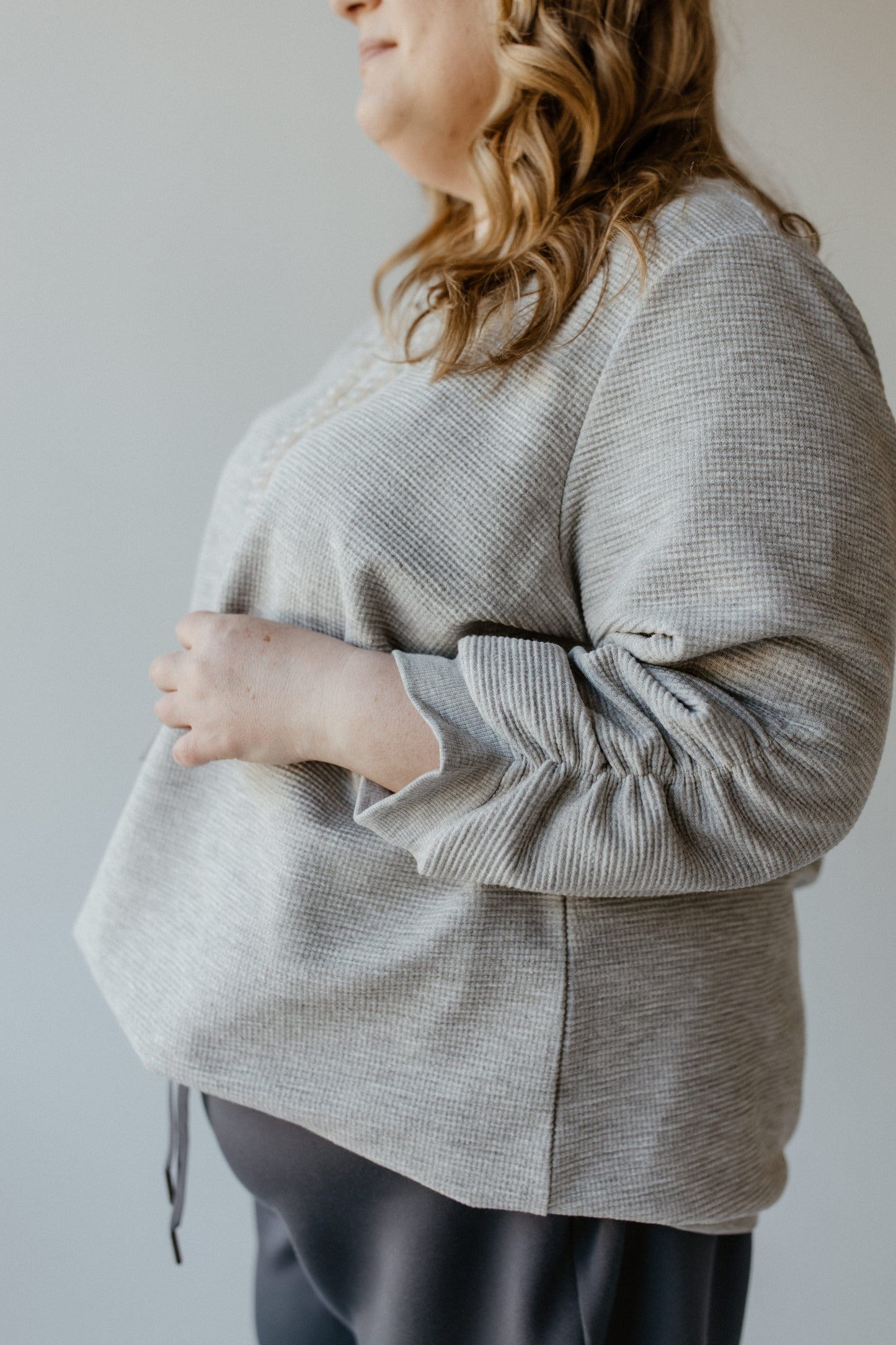 The person is wearing a gray long-sleeve top with ruched sleeves, standing sideways, and gently adjusting the fit. The subtle shimmer of silver lurex adds a touch of elegance to the ensemble.
