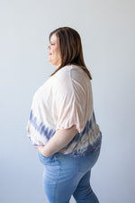 A person with shoulder-length brown hair is seen from behind, wearing the Love Marlow TIE DYE CROPPED TEE IN PEARL, crafted from lightweight fabric in light pink and blue hues, paired with light blue pants against a plain background.