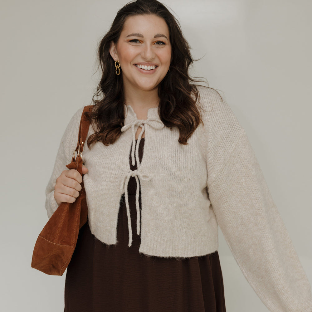 Woman wearing a beige cardigan over a brown dress, holding a brown bag against a white background.