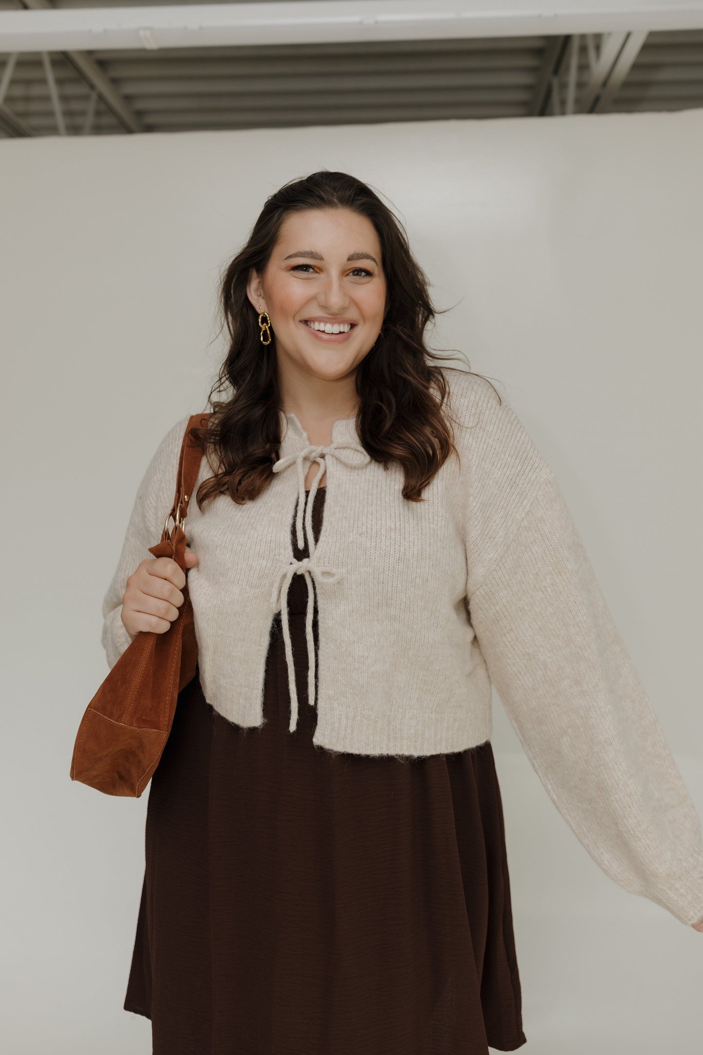 Woman wearing a beige cardigan over a brown dress, holding a brown bag against a white background.