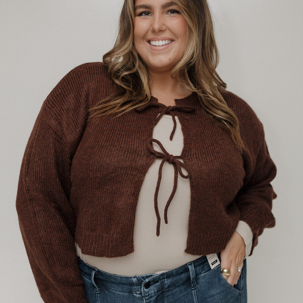 Woman wearing a brown sweater with a bow detail and blue jeans against a plain background