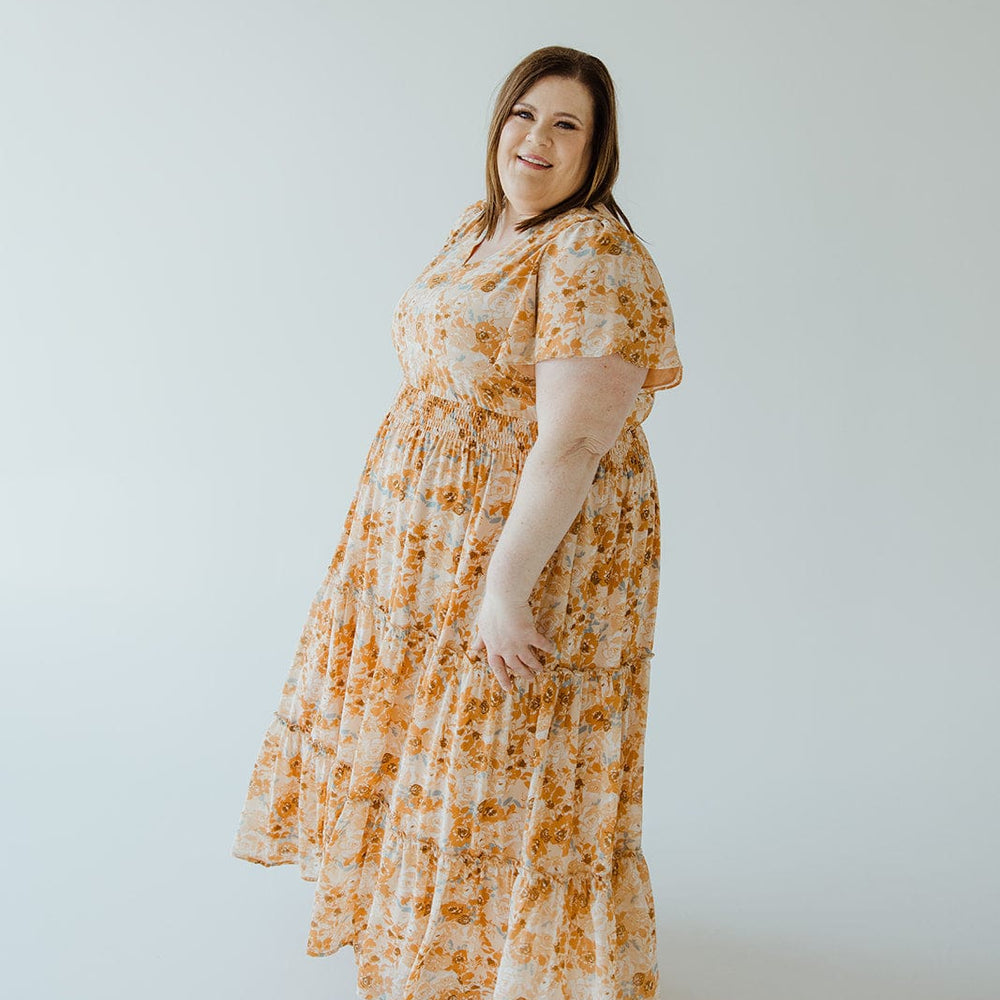 A woman with long hair wears a TIERED FLUTTER SLEEVE FLORAL DRESS IN AMBER SUNSET by Mikarose, featuring a smocked waist, and stands in a light-colored studio.