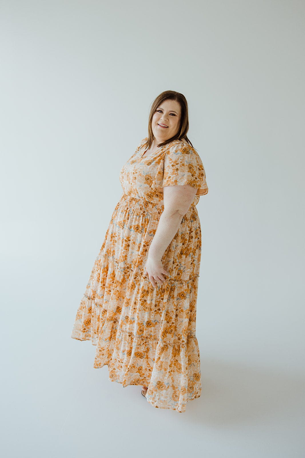 A woman with long hair wears a TIERED FLUTTER SLEEVE FLORAL DRESS IN AMBER SUNSET by Mikarose, featuring a smocked waist, and stands in a light-colored studio.