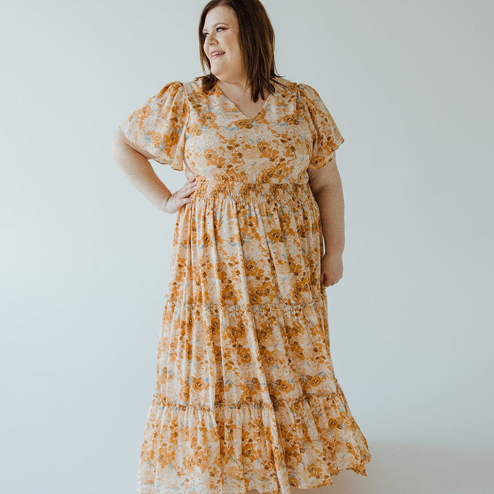 A person standing against a plain background, wearing Mikarose’s TIERED FLUTTER SLEEVE FLORAL DRESS IN AMBER SUNSET. This ankle-length dress features short sleeves and a smocked waist, making it perfect for capturing the essence of an amber sunset.