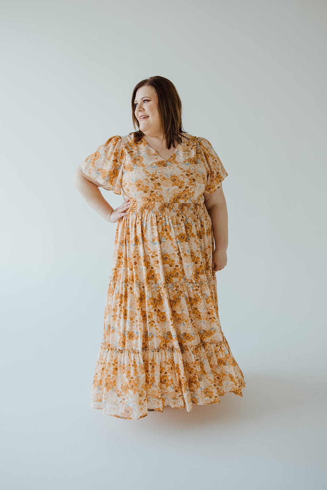 A person standing against a plain background, wearing Mikarose’s TIERED FLUTTER SLEEVE FLORAL DRESS IN AMBER SUNSET. This ankle-length dress features short sleeves and a smocked waist, making it perfect for capturing the essence of an amber sunset.