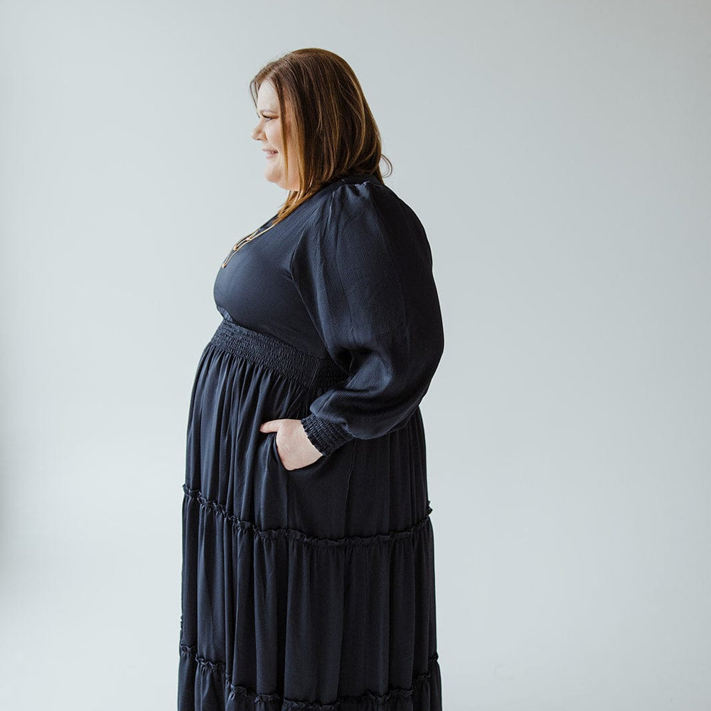 A person in a long, dark party dress with a tiered skirt stands in profile against a light background.