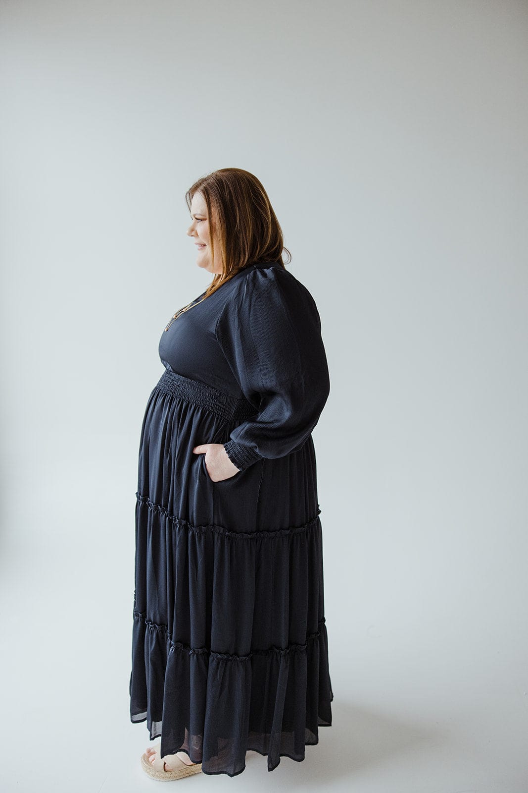 A person in a long, dark party dress with a tiered skirt stands in profile against a light background.